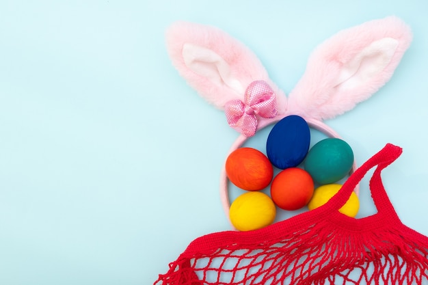 Concetto di acquisto di Pasqua. Uova di Pasqua dipinte a mano, orecchie di coniglietto rosa e una borsa della spesa rossa su sfondo blu, copia spazio, vista dall'alto, piatto lay. Felice Pasqua concetto