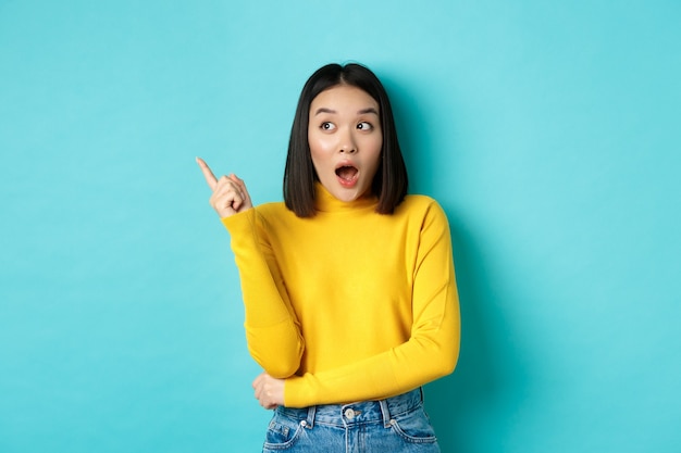 Concetto di acquisto. Ragazza asiatica colpita in pullover giallo, indicando e guardando a sinistra stupita, mostrando la bandiera del logo, in piedi su sfondo blu.