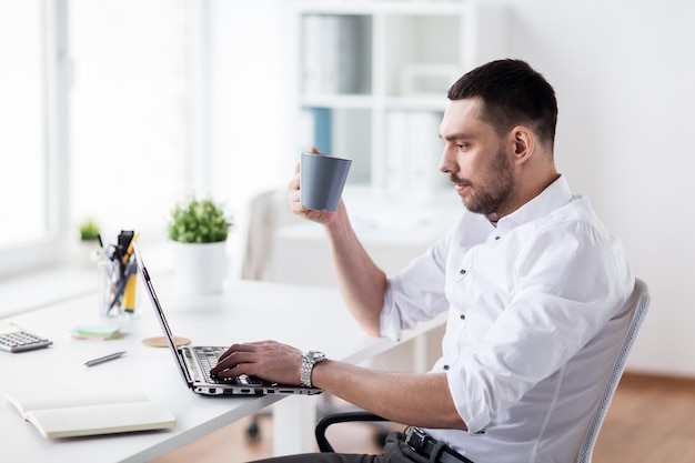 concetto di affari, persone e tecnologia - uomo d'affari che beve caffè e digita sul portatile in ufficio