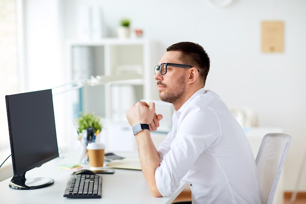 concetto di affari, persone e tecnologia - uomo d'affari con gli occhiali seduto al computer dell'ufficio e pensando