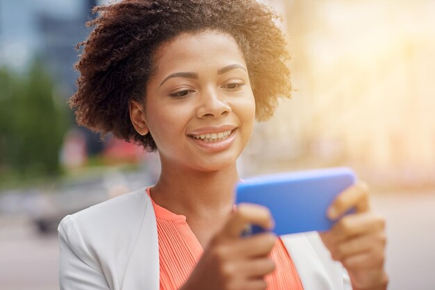 concetto di affari, tecnologia, comunicazione e persone - giovane donna d'affari afroamericana sorridente con smartphone che legge un messaggio sulla strada della città