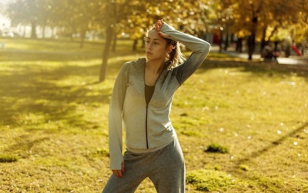 Concetto di allenamento all'aperto. Una donna sportiva stanca in abiti sportivi si asciuga il sudore dalla fronte con la manica nel parco autunnale