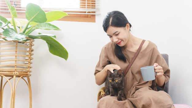 Concetto di amante degli animali La giovane donna asiatica si rilassa bevendo caffè e giocando con il gatto in soggiorno