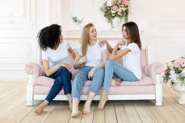 Concetto di amicizia e felicità - tre amiche che parlano a casa. Tre felici e divertenti signore multietniche migliori amiche che ridono divertendosi, donne piuttosto diverse indossano magliette bianche e jeans