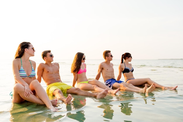 concetto di amicizia, mare, vacanze estive, vacanze e persone - gruppo di amici sorridenti che indossano costumi da bagno e occhiali da sole seduti in acqua sulla spiaggia