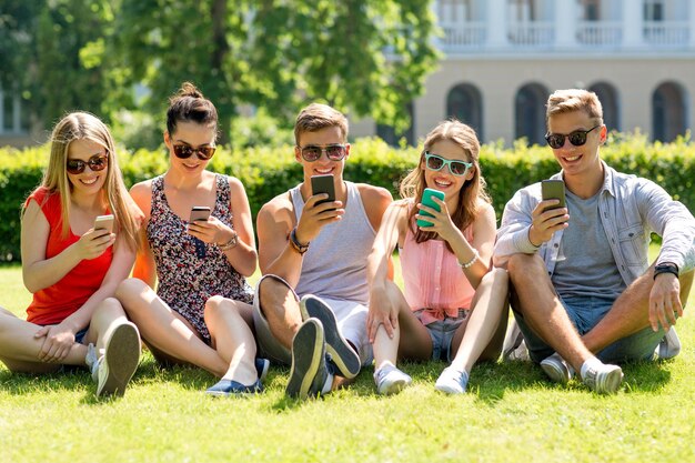 concetto di amicizia, tempo libero, estate, tecnologia e persone - gruppo di amici sorridenti con smartphone seduti sull'erba nel parco