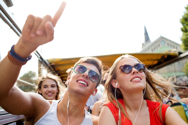 concetto di amicizia, viaggi, vacanze, estate e persone - coppia sorridente con auricolari che viaggiano in bus turistico e dito puntato