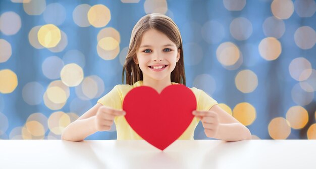 concetto di amore, carità, vacanze, bambini e persone - bambina sorridente con cuore rosso su sfondo di luci blu