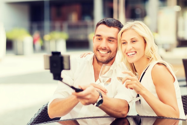 concetto di amore, data, tecnologia, persone e vacanze - coppia felice felice che scatta foto con lo smartphone sul bastone per selfie e bicchieri tintinnanti al caffè o al ristorante della strada della città