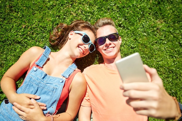 concetto di amore e persone - felice coppia adolescente in occhiali da sole sdraiati sull'erba e prendendo selfie sullo smartphone in estate