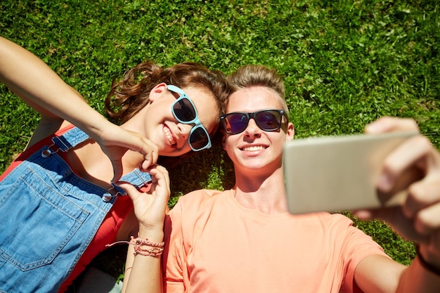 concetto di amore e persone - felice coppia adolescente in occhiali da sole sdraiati sull'erba e prendendo selfie sullo smartphone in estate