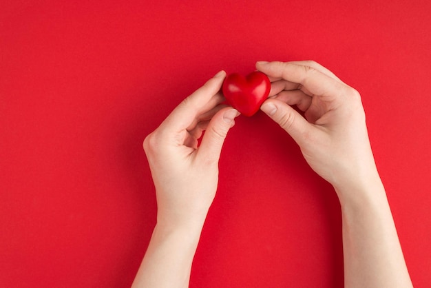 Concetto di amore e romanticismo. In alto sopra la foto vista dall'alto delle mani femminili che tengono il cuore rosso isolato su sfondo rosso con copyspace