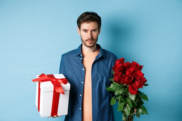 Concetto di amore e San Valentino. Uomo appassionato che guarda fiducioso alla macchina fotografica, che tiene scatola regalo e rose rosse per un appuntamento romantico, in piedi su sfondo blu.