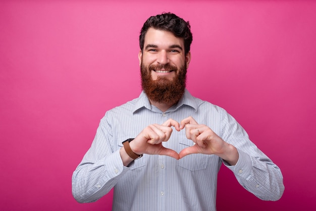 Concetto di amore. Giovane uomo barbuto che sorride alla telecamera facendo un gesto di cuore.