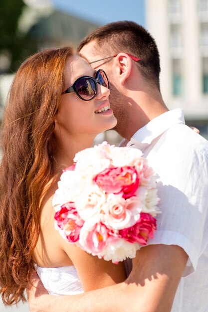 concetto di amore, matrimonio, estate, appuntamenti e persone - coppia sorridente che indossa occhiali da sole con un mazzo di fiori che si abbracciano in città