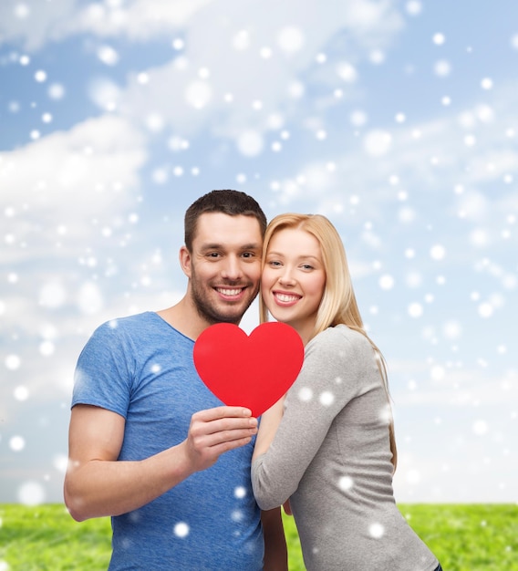 concetto di amore, persone e famiglia - coppia sorridente con forma di cuore di carta rossa che abbraccia su sfondo blu cielo, neve ed erba