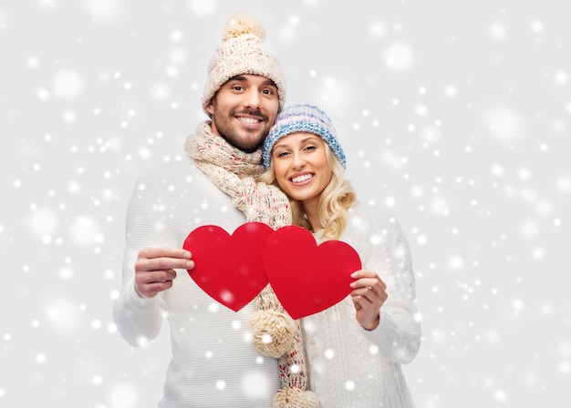 concetto di amore, san valentino, coppia, natale e persone - uomo e donna sorridenti in cappelli invernali e sciarpa che tengono forme di cuore di carta rossa
