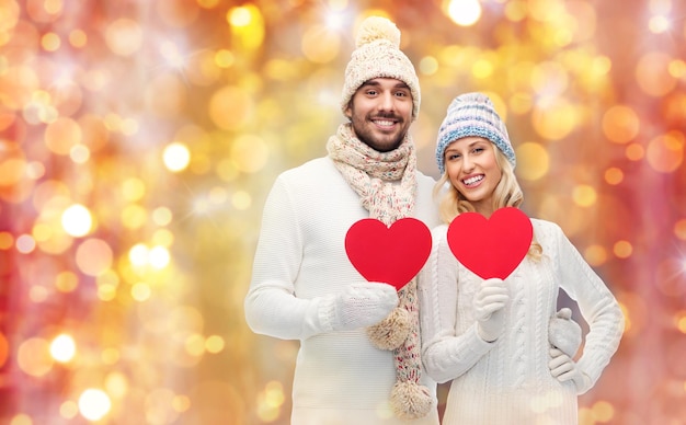 concetto di amore, San Valentino, coppia, Natale e persone - uomo e donna sorridenti in cappelli invernali e sciarpa con forme di cuore di carta rossa su sfondo di luci