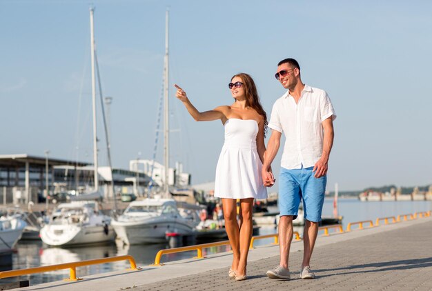 concetto di amore, viaggi, turismo e persone - coppia sorridente che indossa occhiali da sole che cammina al porto e punta il dito