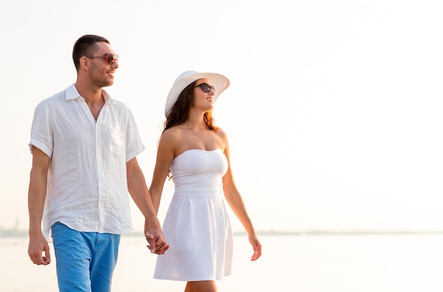 concetto di amore, viaggi, turismo, estate e persone - coppia sorridente in vacanza che indossa occhiali da sole e si tiene per mano camminando in riva al mare