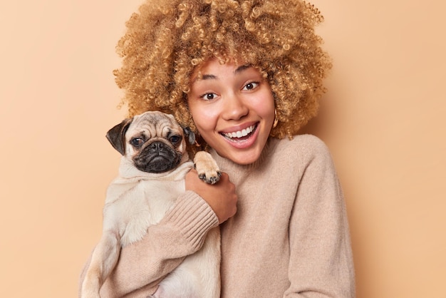 Concetto di animali domestici e persone. La donna gioiosa con i capelli ricci posa con il cane carino abbraccia l'animale domestico sorride felicemente vestito casualmente isolato su sfondo beige abbraccia il nuovo adorabile membro della famiglia