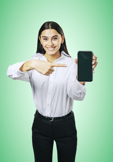 Concetto di applicazione mobile con ragazza felice in camicia bianca che mostra smartphone moderno con schermo vuoto mockup su sfondo verde astratto