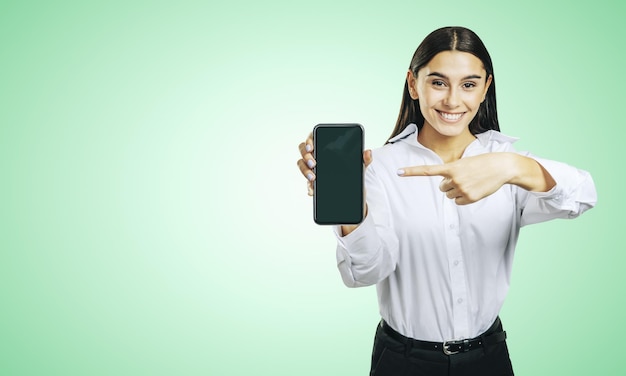 Concetto di applicazione mobile con ragazza felice in camicia bianca che mostra uno smartphone moderno con schermo vuoto su sfondo verde chiaro astratto con posto per il tuo logo o testo mock up