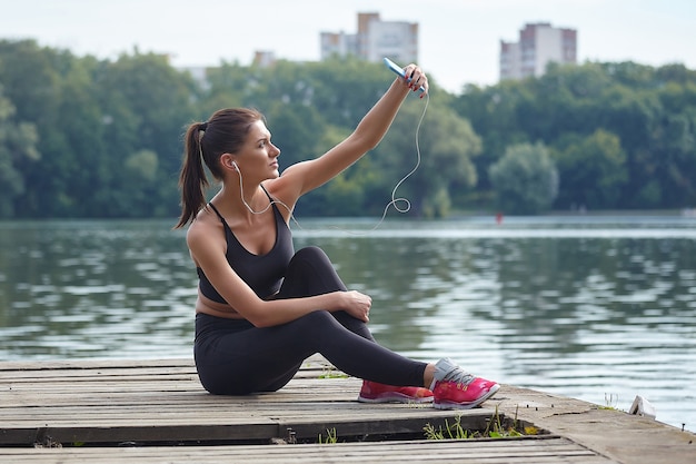 Concetto di apprendimento a distanza. Attraente ragazza in abiti sportivi si siede su un molo in legno in un parco cittadino. Lezioni online tramite smartphone.