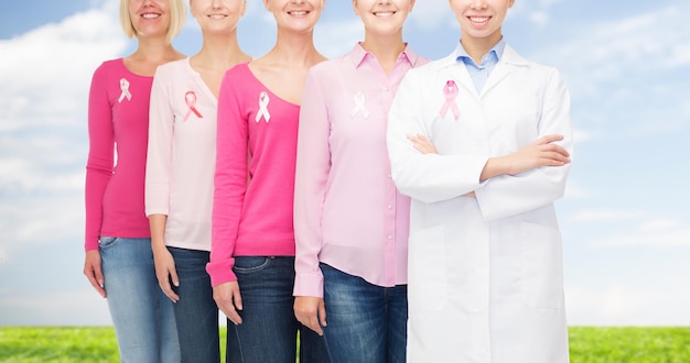 concetto di assistenza sanitaria, persone e medicina - primo piano di donne sorridenti in camicie bianche con nastri rosa per la consapevolezza del cancro al seno su sfondo blu cielo e erba