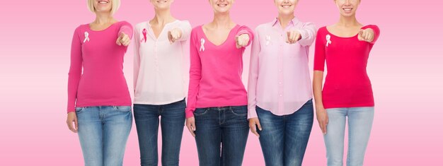 concetto di assistenza sanitaria, persone, gesto e medicina - primo piano di donne sorridenti in camicie bianche con nastri di consapevolezza del cancro al seno che puntano su di te su sfondo rosa
