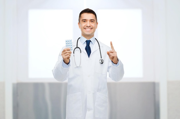 concetto di assistenza sanitaria, professione, persone e medicina - medico maschio sorridente in camice bianco con pillole che puntano il dito verso l'alto