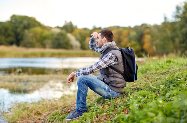 concetto di avventura, viaggi, turismo, escursione e persone - uomo sorridente con zaino che riposa sulla riva del fiume