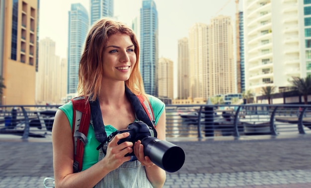 concetto di avventura, viaggio, turismo, escursione e persone - giovane donna felice con zaino e macchina fotografica che fotografa sulla città di dubai e sul porto con sfondo di barche