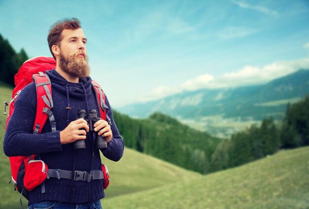 concetto di avventura, viaggio, turismo, escursione e persone - uomo con zaino rosso e binocolo su sfondo di colline alpine