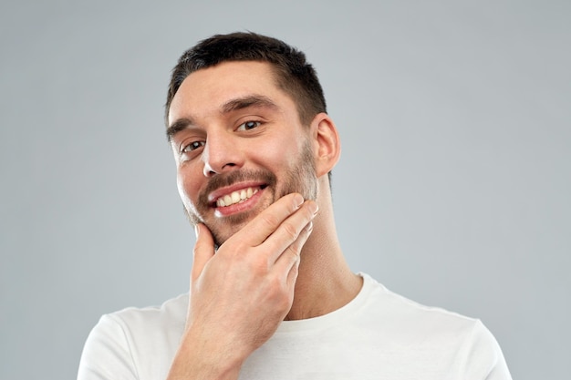 concetto di bellezza, cura della pelle, toelettatura e persone - giovane felice che tocca il viso o la barba su sfondo grigio