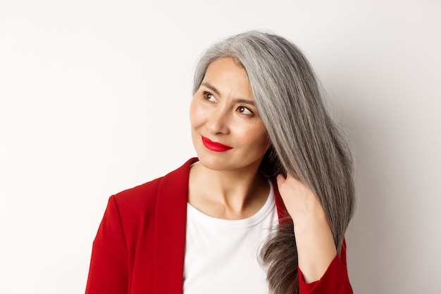 Concetto di bellezza e cura dei capelli. Primo piano di un'elegante donna anziana asiatica che mostra capelli grigi lucenti e sani, sorridendo e guardando da parte, sfondo bianco