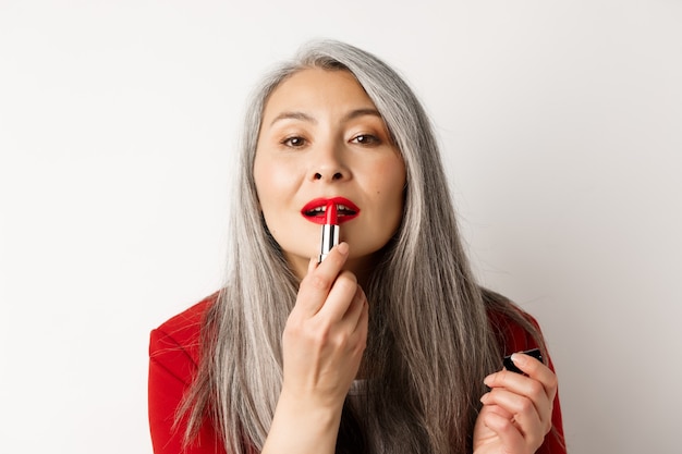 Concetto di bellezza e moda. Primo piano di una donna asiatica anziana che si guarda allo specchio e applica il rossetto rosso, in piedi su sfondo bianco