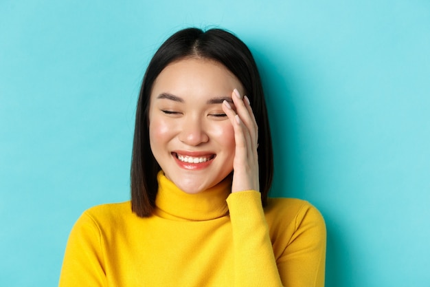 Concetto di bellezza e trucco. Primo piano di bella donna asiatica ridendo e guardando arrossato dai complimenti, in piedi su sfondo blu.