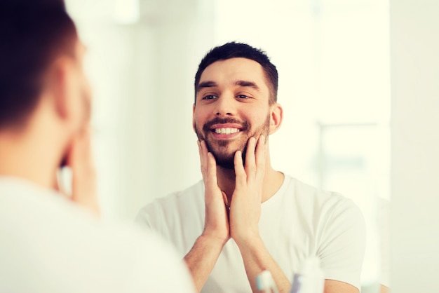 concetto di bellezza, igiene e persone - giovane sorridente che guarda allo specchio nel bagno di casa