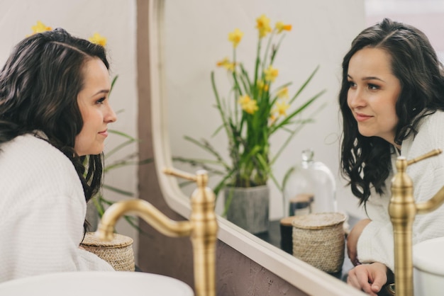 Concetto di bellezza, igiene, mattina e persone - giovane donna che guarda allo specchio nel bagno di casa