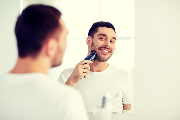 concetto di bellezza, igiene, rasatura, toelettatura e persone - giovane che cerca di specchiarsi e radersi la barba con il rifinitore o il rasoio elettrico nel bagno di casa