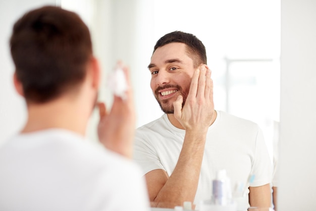 concetto di bellezza, igiene, rasatura, toelettatura e persone - giovane sorridente che guarda allo specchio e applica la schiuma da barba sul viso nel bagno di casa