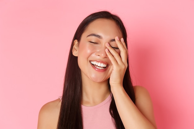 Concetto di bellezza, moda e stile di vita. Primo piano di una bella ragazza asiatica senza acne o macchie, sorriso bianco, viso toccante e che sembra felice, in piedi sopra il muro rosa.