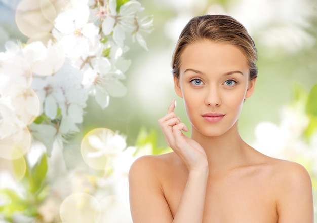 concetto di bellezza, persone, cosmetici, cura della pelle e salute - giovane donna che applica la crema sul viso su sfondo verde naturale con fiori di ciliegio