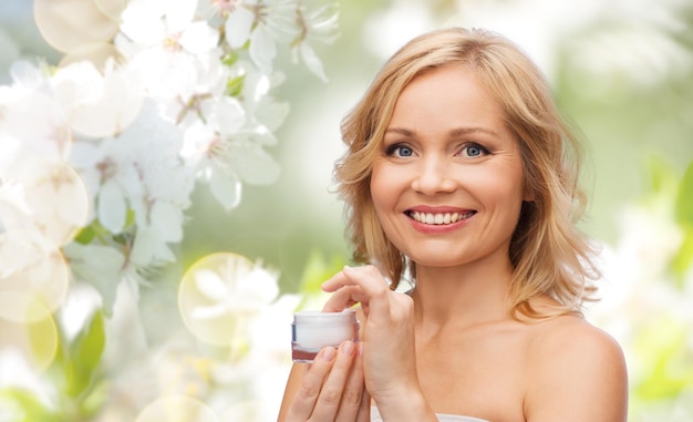 concetto di bellezza, persone, cura della pelle e cosmetici - donna felice con vasetto di crema su sfondo di fiori di ciliegio primaverile naturale