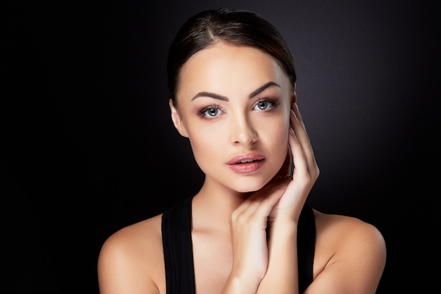 Concetto di bellezza, ritratto di testa e spalle della bellissima modella con trucco nudo che guarda l'obbiettivo, toccando il viso. Donna castana in studio con sfondo nero