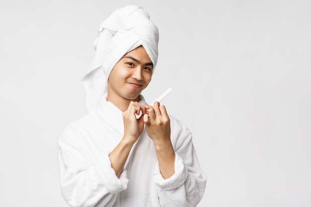 Concetto di bellezza, spa e tempo libero. Ritratto di sciocco bell'uomo asiatico in accappatoio e telo da bagno, sorridendo compiaciuto, lucidando le unghie e fare la manicure, in piedi muro bianco