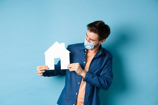 Concetto di bene immobile e quarantena. Giovane uomo caucasico nella mascherina medica guardando il ritaglio di casa di carta, in piedi su sfondo blu.