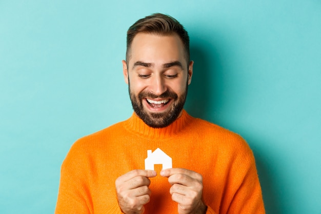 Concetto di bene immobile. Felice giovane uomo alla ricerca di una casa in affitto, tenendo in mano carta maket casa e sorridente, in piedi su sfondo blu.