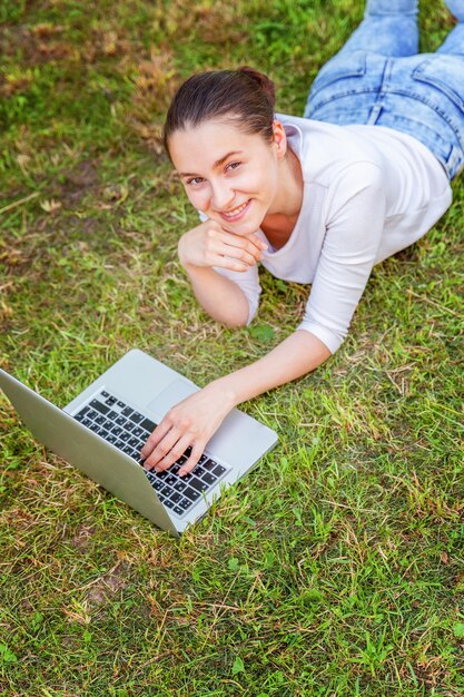 Concetto di business freelance. Giovane donna sdraiata sul prato di erba verde nel parco cittadino che lavora su un computer portatile. Stile di vita autentico studentessa candida che studia all'aperto. Ufficio Mobile